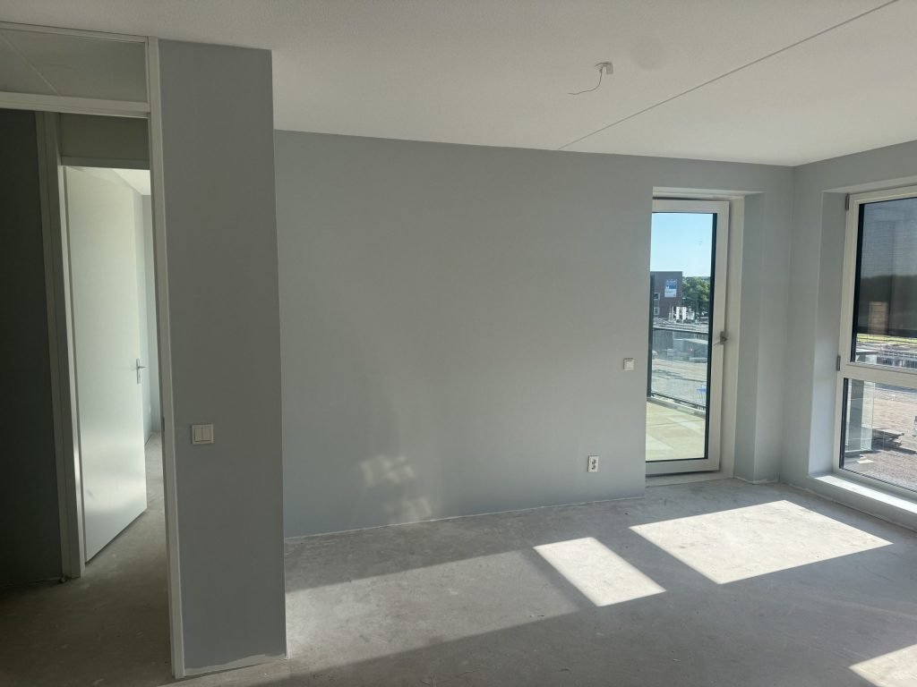 Nieuw appartement met balkondeur en uitzicht op een bouwterrein, de wanden egaal lichtgrijs geverfd