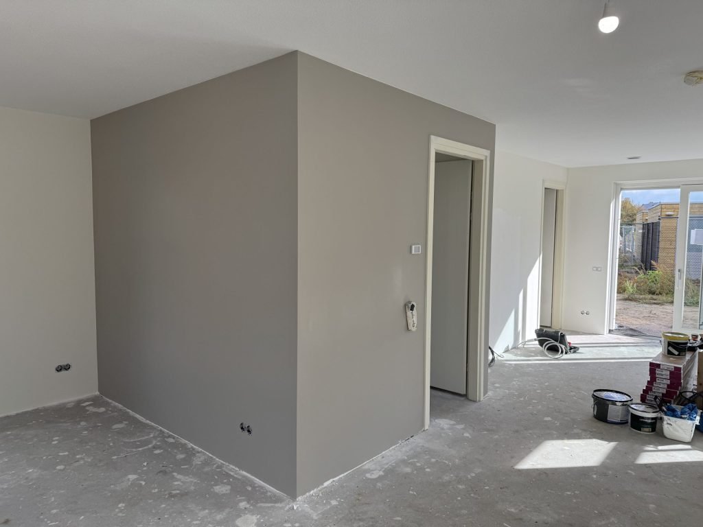 Open woonruimte in een nieuwbouwwoning met een grijs geverfde vlieswand en verfemmers op de vloer